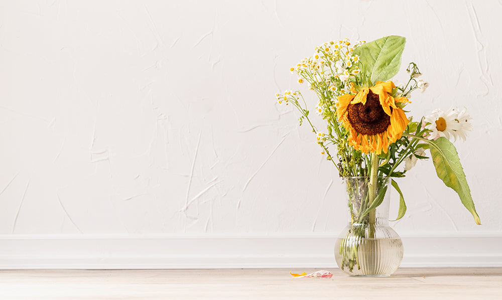Wilting flowers in a vase, including a drooping sunflower and daisies