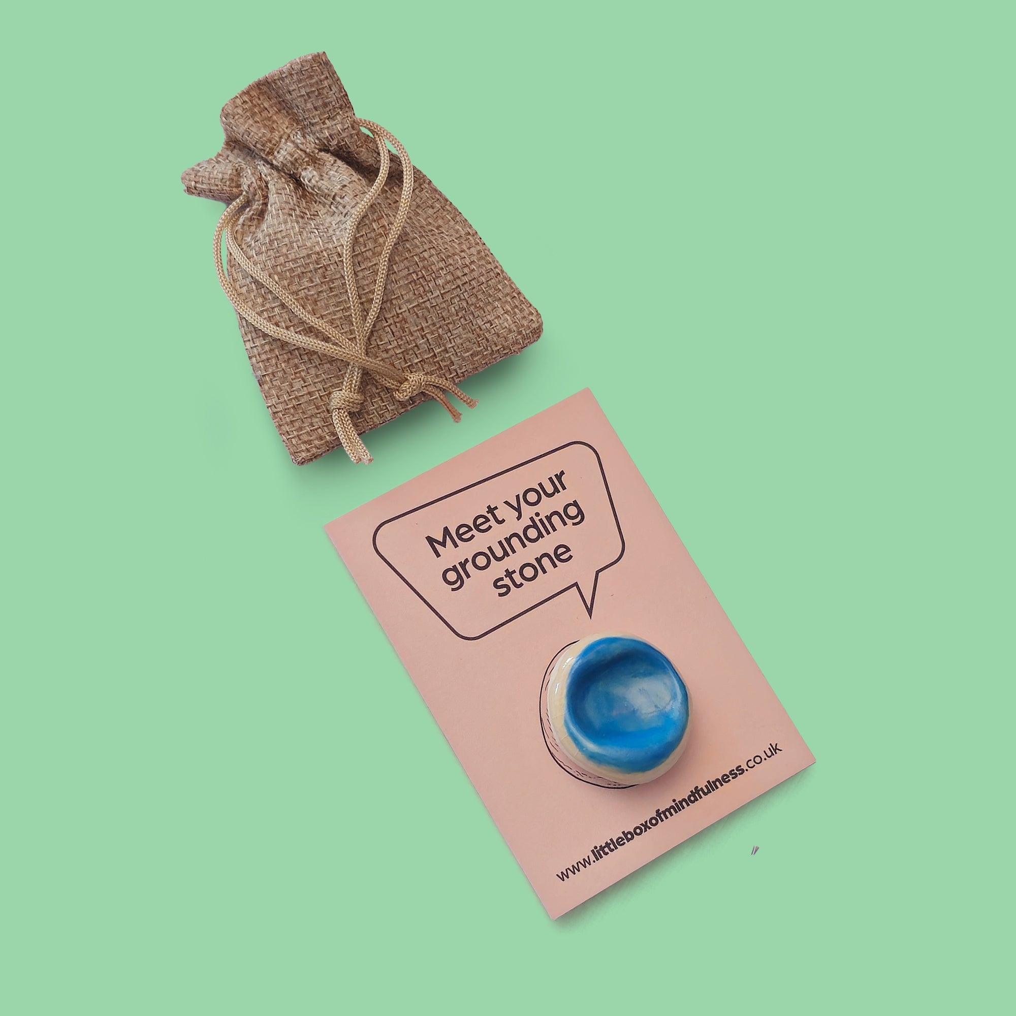 A small blue grounding stone displayed on a card that reads 'Meet your grounding stone,' with a small burlap drawstring pouch above on a solid mint green background.