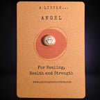 A pocket pal with angel illustration sits centered on a kraft paper backing card. The card has printed text reading 'A LITTLE ANGEL'