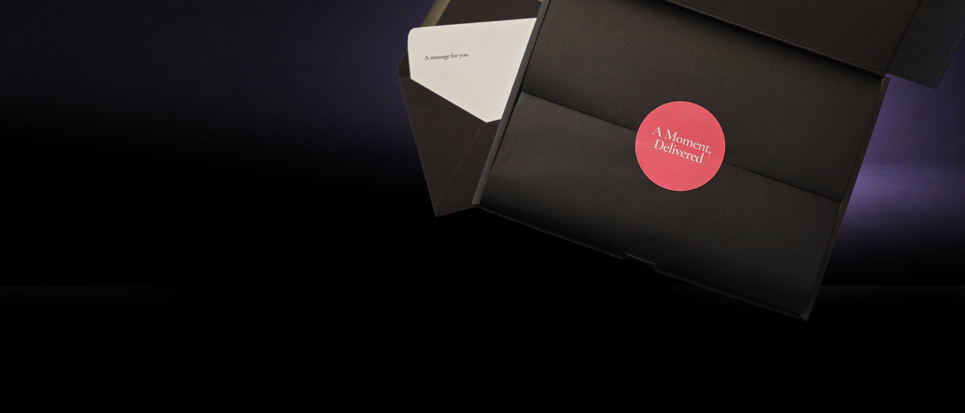 A black gift box with a white envelope partially visible inside, featuring a circular red sticker that reads 'A Moment of Time' on a dark background.