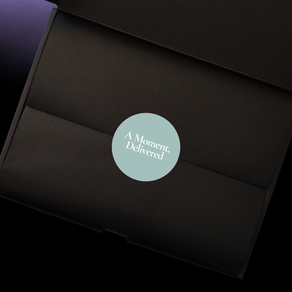 A close-up of a black gift box interior with a circular blue grey sticker in the centre that reads 'A Moment, Delivered' in white text.