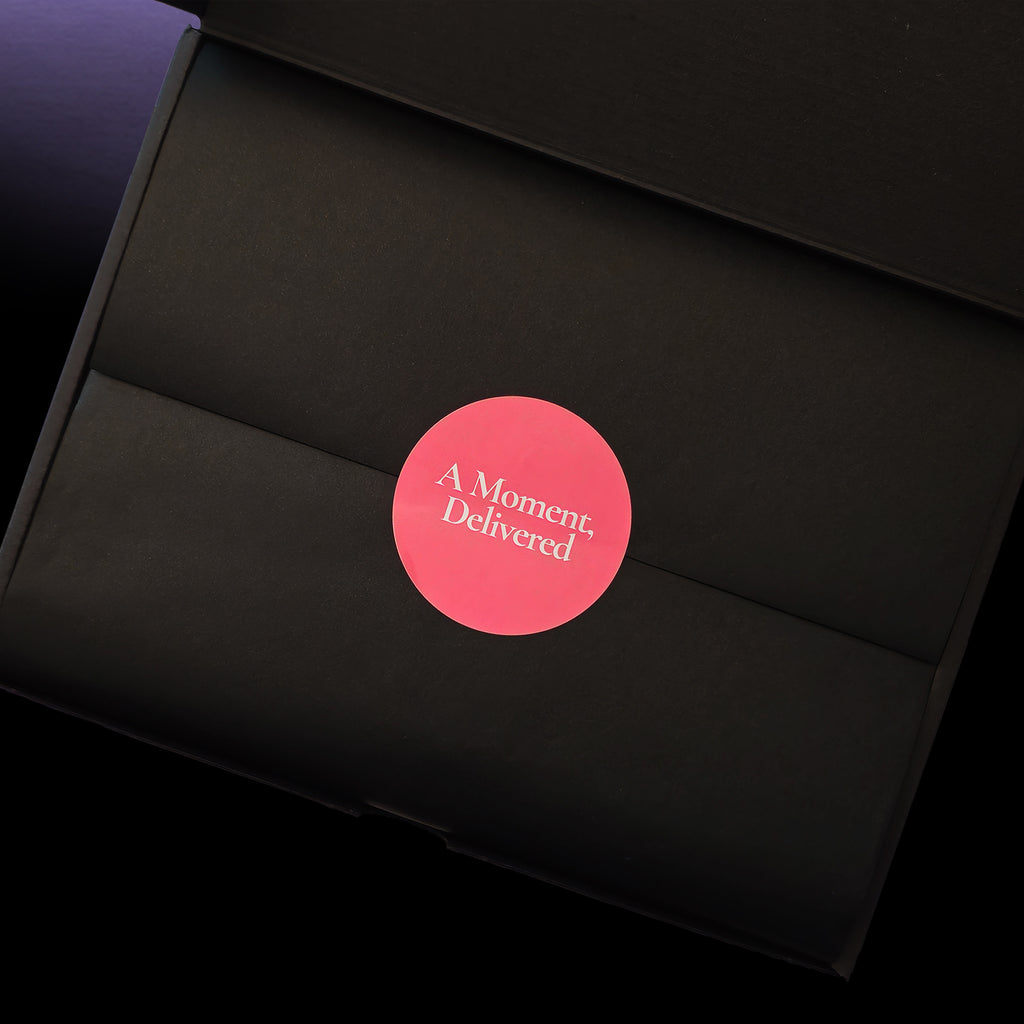 A close-up of a black gift box with a circular pink sticker in the center that reads 'A Moment, Delivered' in white text.