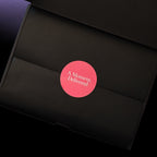 A close-up of a black gift box with a circular pink sticker in the center that reads 'A Moment, Delivered' in white text.