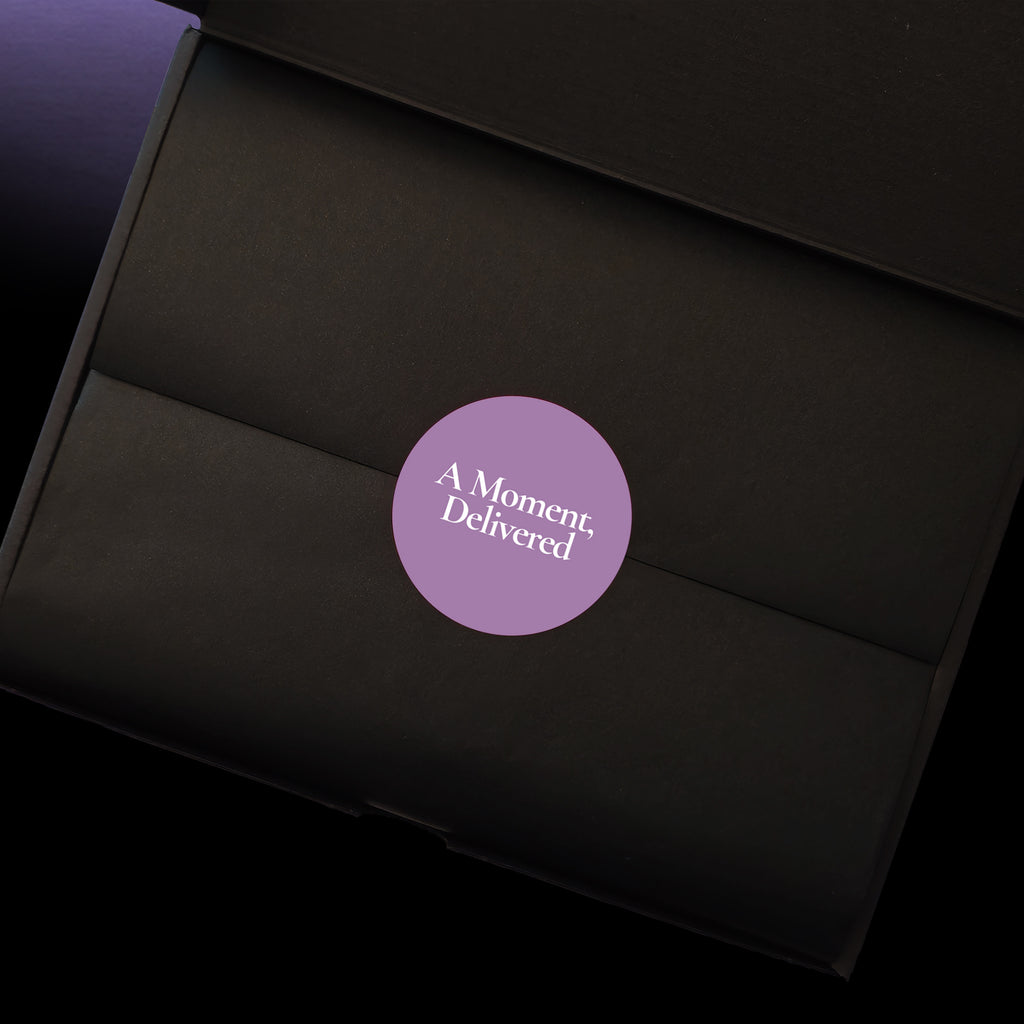 A close-up of a black gift box interior with a circular purple sticker in the centre that reads 'A Moment, Delivered' in white text.