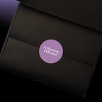 A close-up of a black gift box interior with a circular purple sticker in the centre that reads 'A Moment, Delivered' in white text.