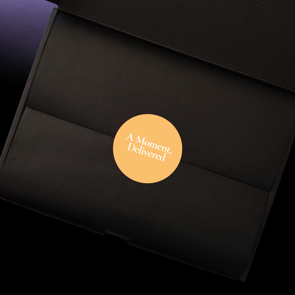 A close-up of a black gift box interior with a circular yellow sticker in the cente that reads 'A Moment, Delivered' in white text.
