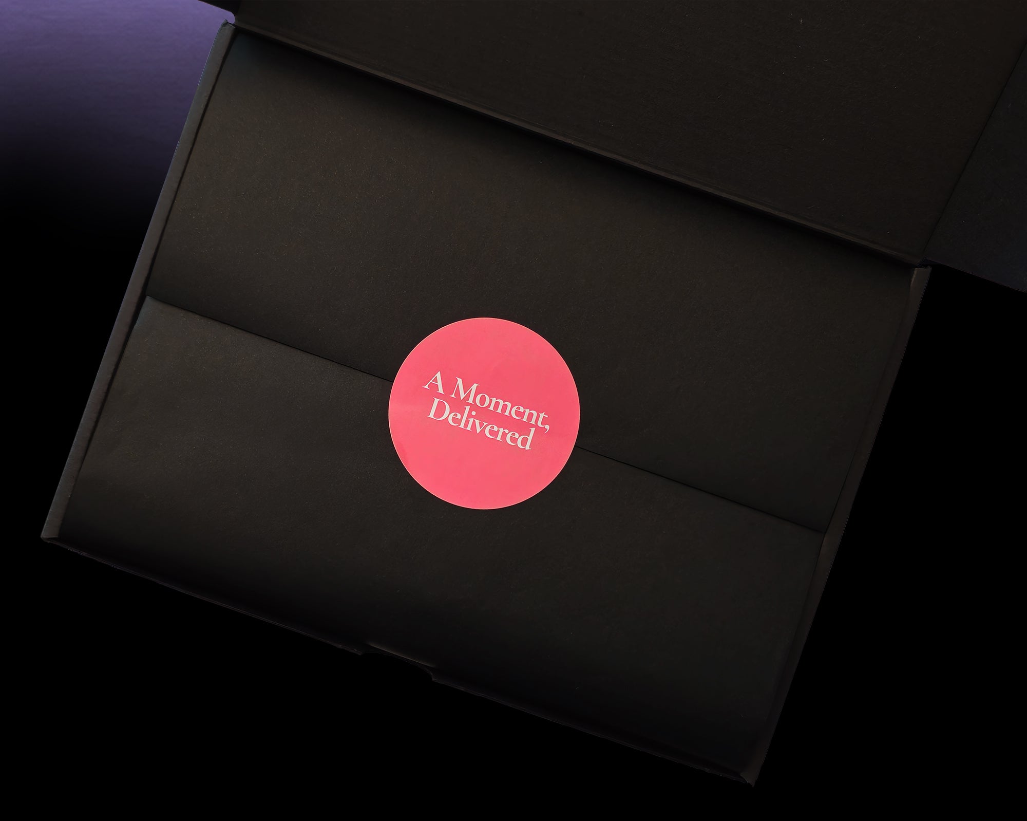 A black presentation box with its lid open, featuring a circular pink sticker in the center with the white text 'A Moment. Delivered.