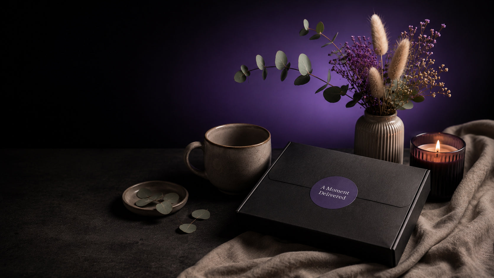 A black gift box with a purple circular label reading 'A Moment Before' sits on a draped fabric surface beside a ceramic mug, a small dish with round items, and a lit candle. A vase with dried flowers and greenery stands in the background against a dark purple-lit backdrop.