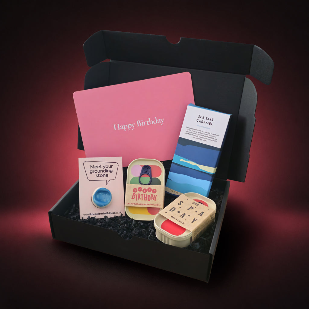 An open black gift box filled with birthday-themed items, including a pink card that says 'Happy Birthday,' a blue and white 'SEA SALT CARAMEL' chocolate bar, a small sign reading 'Meet your grounding stone,' two colourful tins all set against a dark pink gradient background.
