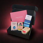 An open black gift box filled with birthday-themed items, including a pink card that says 'Happy Birthday,' a blue and white 'SEA SALT CARAMEL' chocolate bar, a small sign reading 'Meet your grounding stone,' two colourful tins all set against a dark pink gradient background.