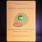 A small green frog pebble with white eyes and a smile is displayed on a kraft paper card