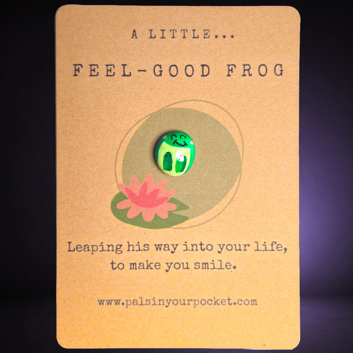 A small green frog pebble with white eyes and a smile is displayed on a kraft paper card