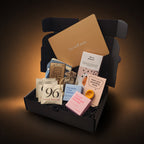 A curated gift box set in an open black box with shredded black filler, featuring a large tan card reading 'Get well soon,' a chocolate bar called 'tea and biscuit', a pastel note card, several tea bags, a small pouch, and other small gift items, photographed on a dark orange background with warm light around the box.