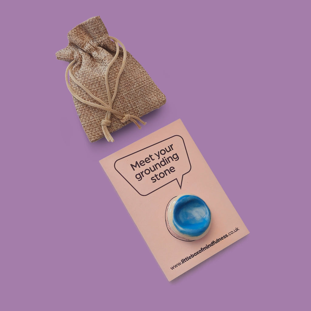 A small blue grounding stone displayed on a card that reads 'Meet your grounding stone,' with a small burlap drawstring pouch above on a solid purple background.