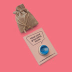 A blue and white circular stone, displayed on a beige card that reads 'Meet your grounding stone' with a website URL. A small beige burlap drawstring pouch is placed above the card on a solid pink background.