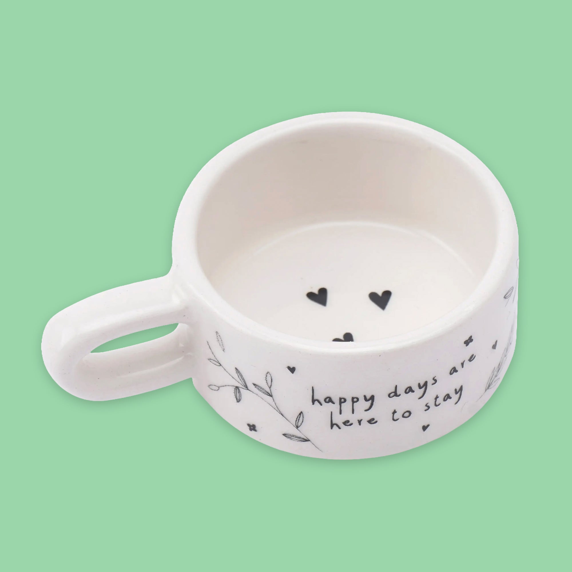 A white ceramic tealight holder with a handle, decorated with black hand-drawn hearts, leaves, and the phrase 'happy days are here to stay' on a light green background.