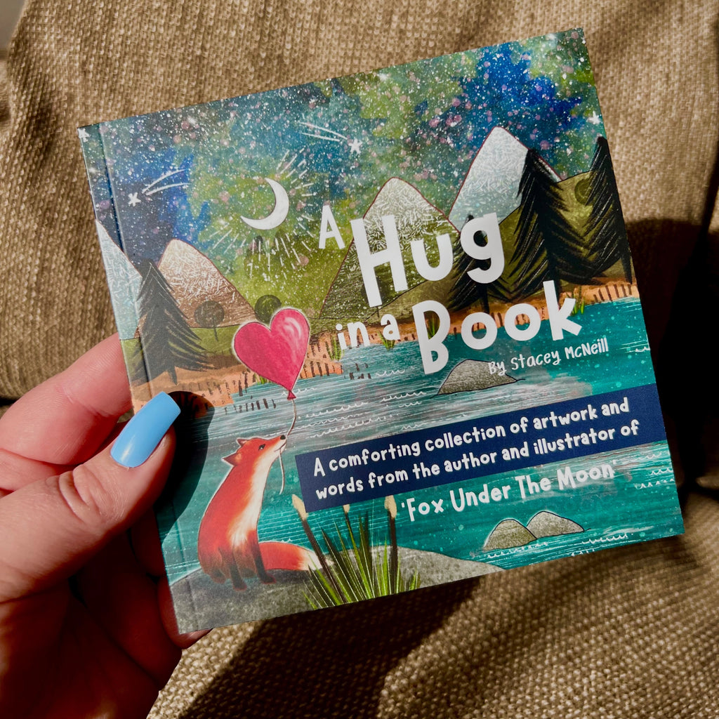 A hand with blue nail polish holds a book titled "A Hug in a Book" by Stacey McNeill. The cover features a colourful illustrated scene with mountains, stars, a crescent moon, a fox, and a pink heart balloon, set against a brown textured fabric background.