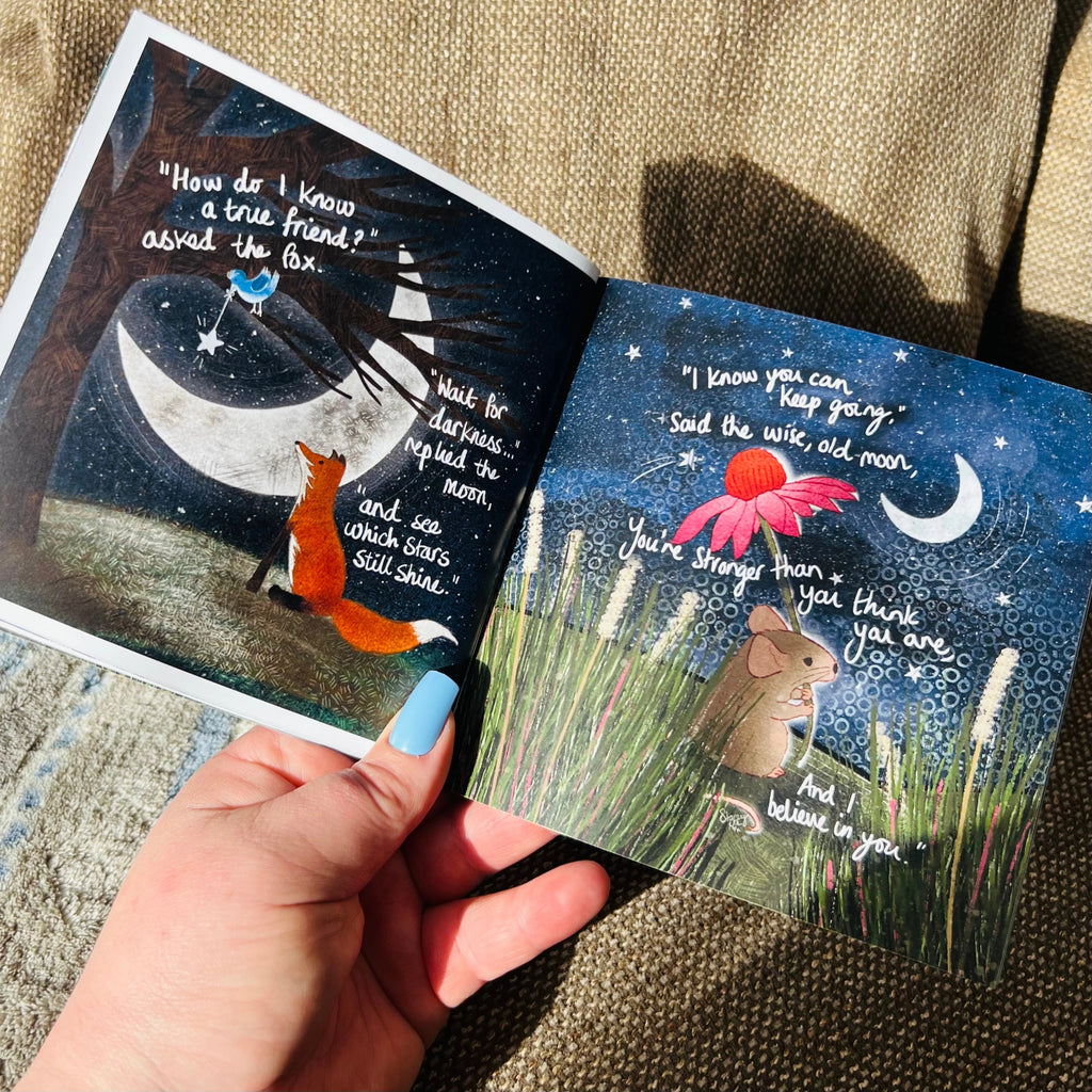 Open book with illustrations of a fox and mouse, featuring text, held by a hand.