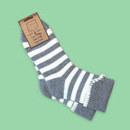 A pair of grey and white striped cuff socks, attached to a brown paper tag that reads 'Jess Lou Super Fluffy, Super Soft Cosy Socks', set against a solid light green background.