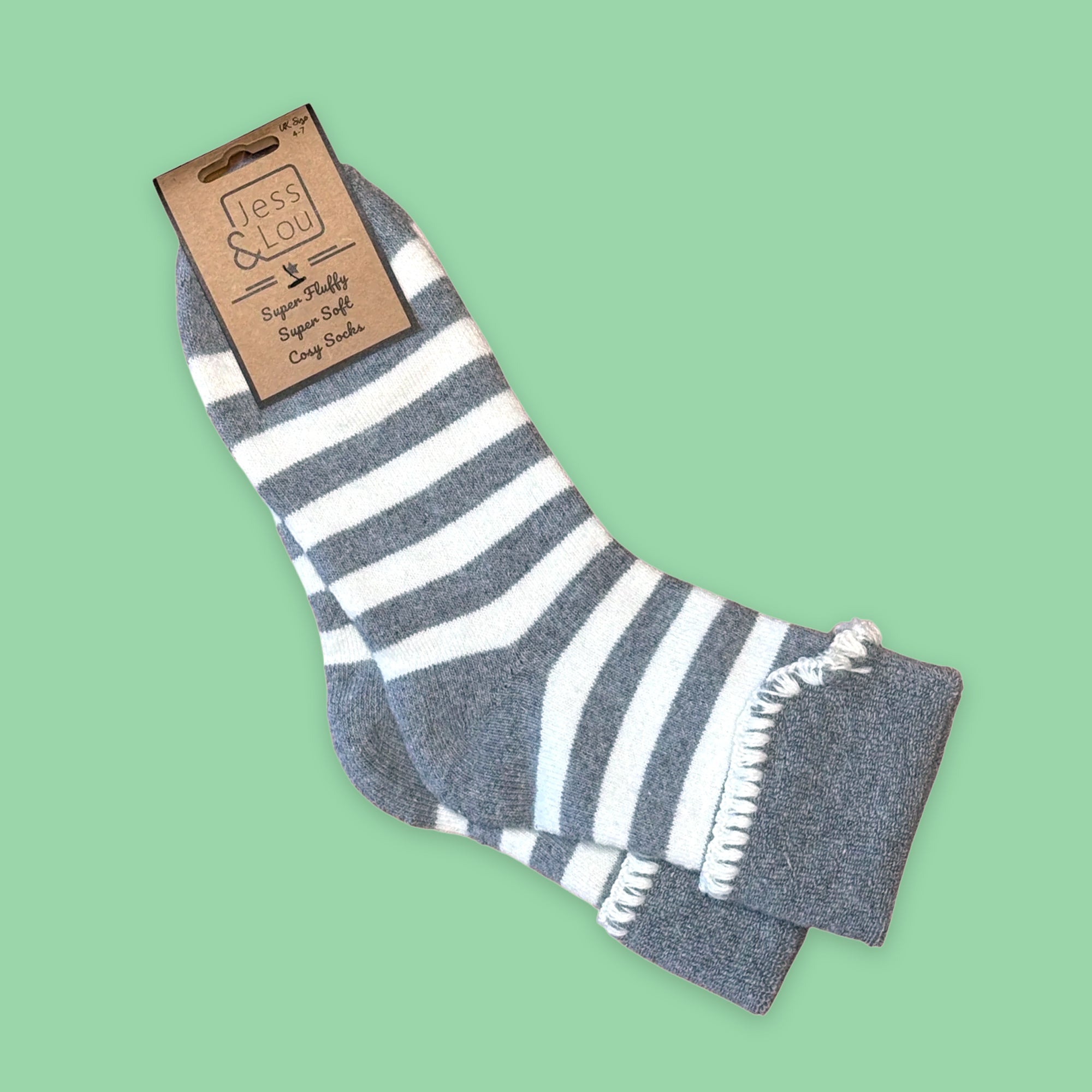 A pair of grey and white striped cuff socks, attached to a brown paper tag that reads 'Jess Lou Super Fluffy, Super Soft Cosy Socks', set against a solid light green background.