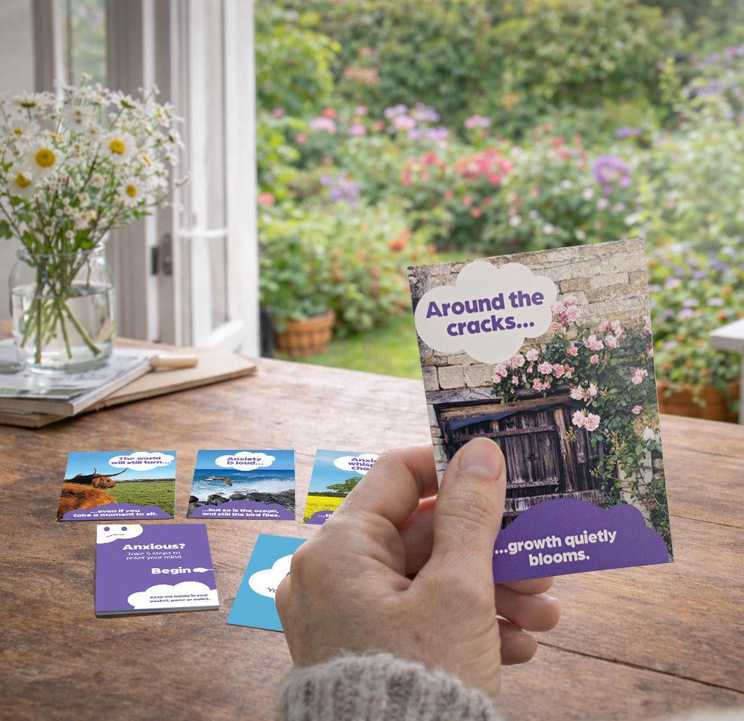 Hand holding affirmation card with inspirational text, surrounded by more cards on a wooden table with a garden view.