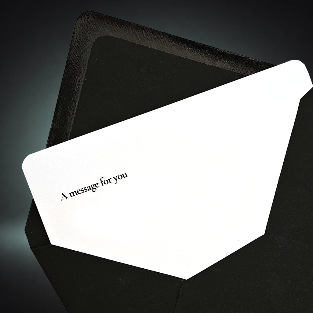 A white greeting card with the printed text 'A message for you' in black, partially tucked inside a textured black envelope against a dark gradient background.