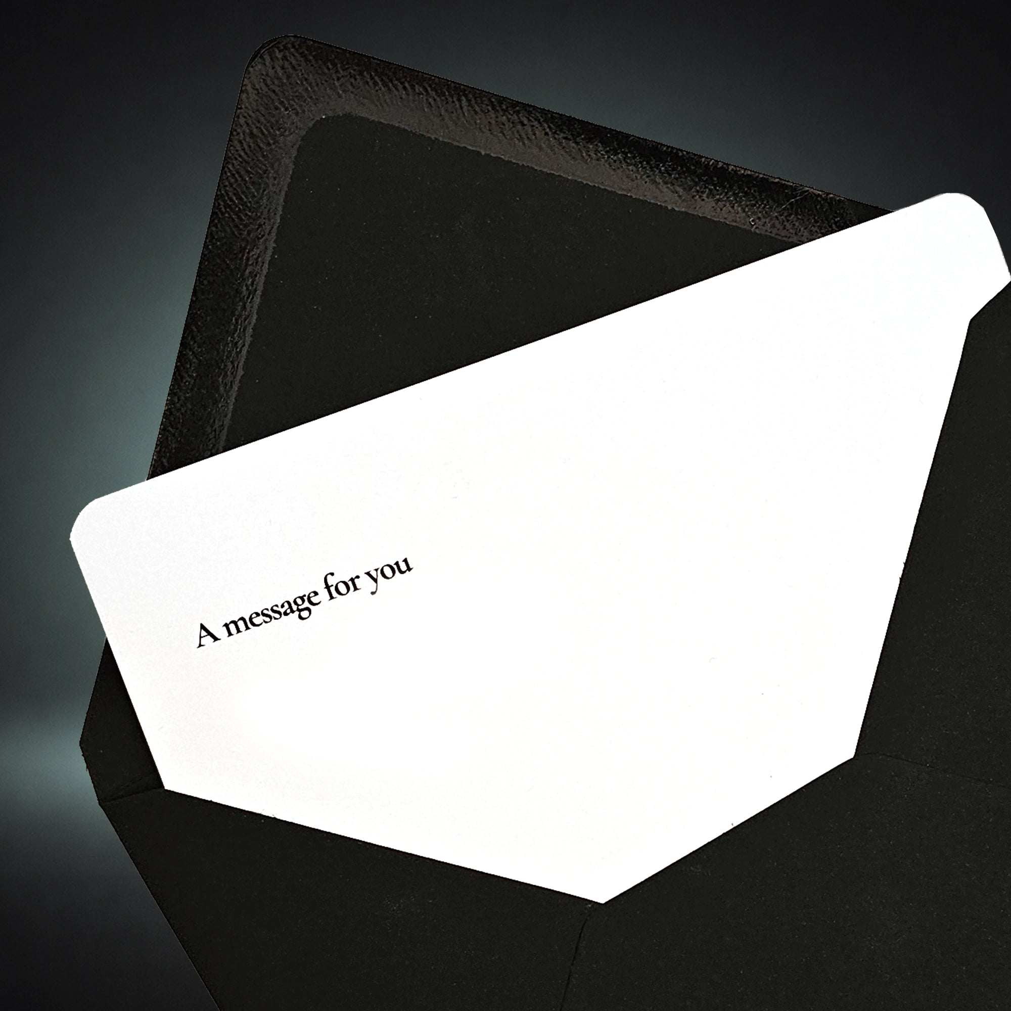 A white greeting card with the printed text 'A message for you' in black, partially tucked inside a textured black envelope against a dark gradient background.
