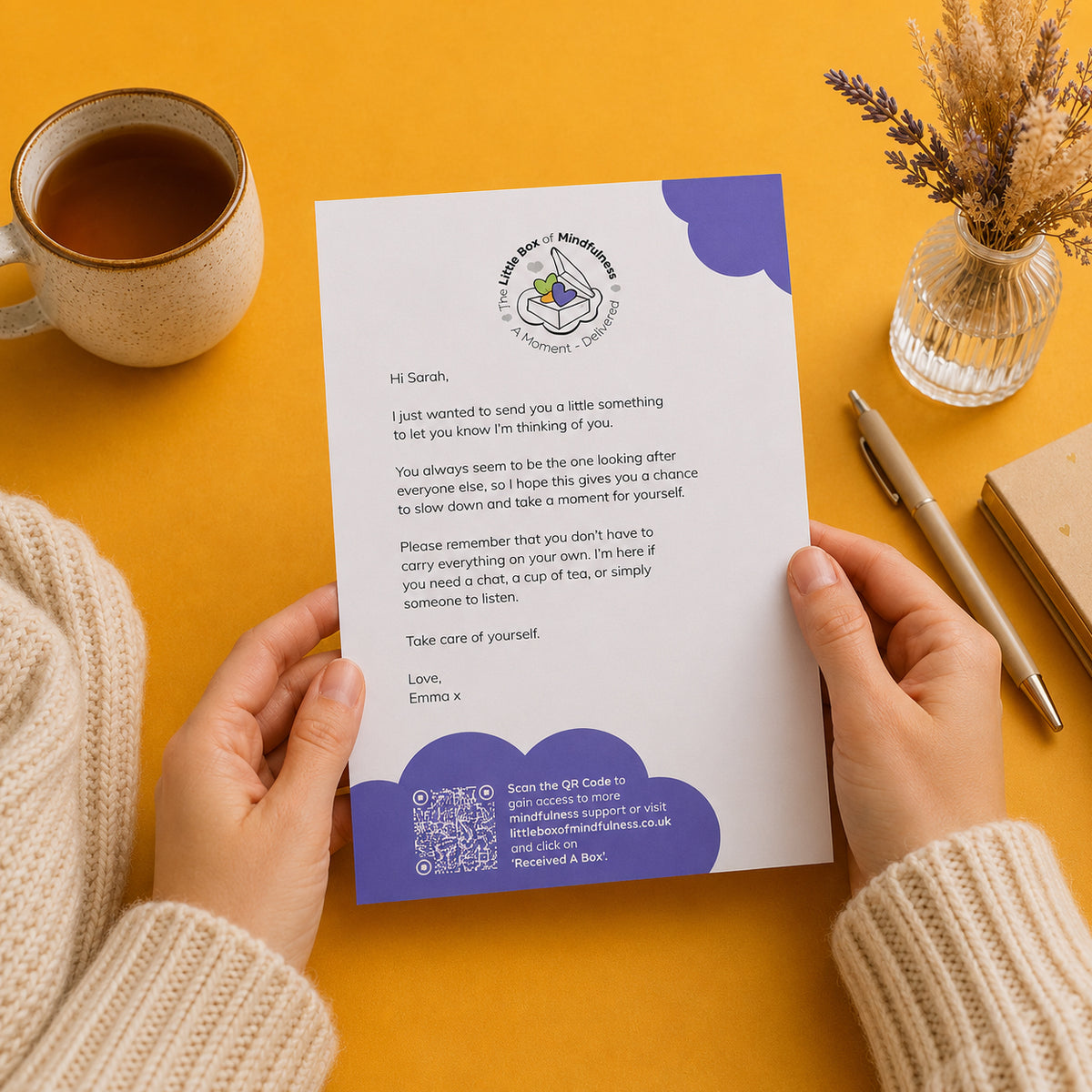 A person holds a personalised letter with a purple and white design, photographed from above on a warm yellow surface. A cup of tea, a small vase with dried stems, and a notebook are arranged around it.