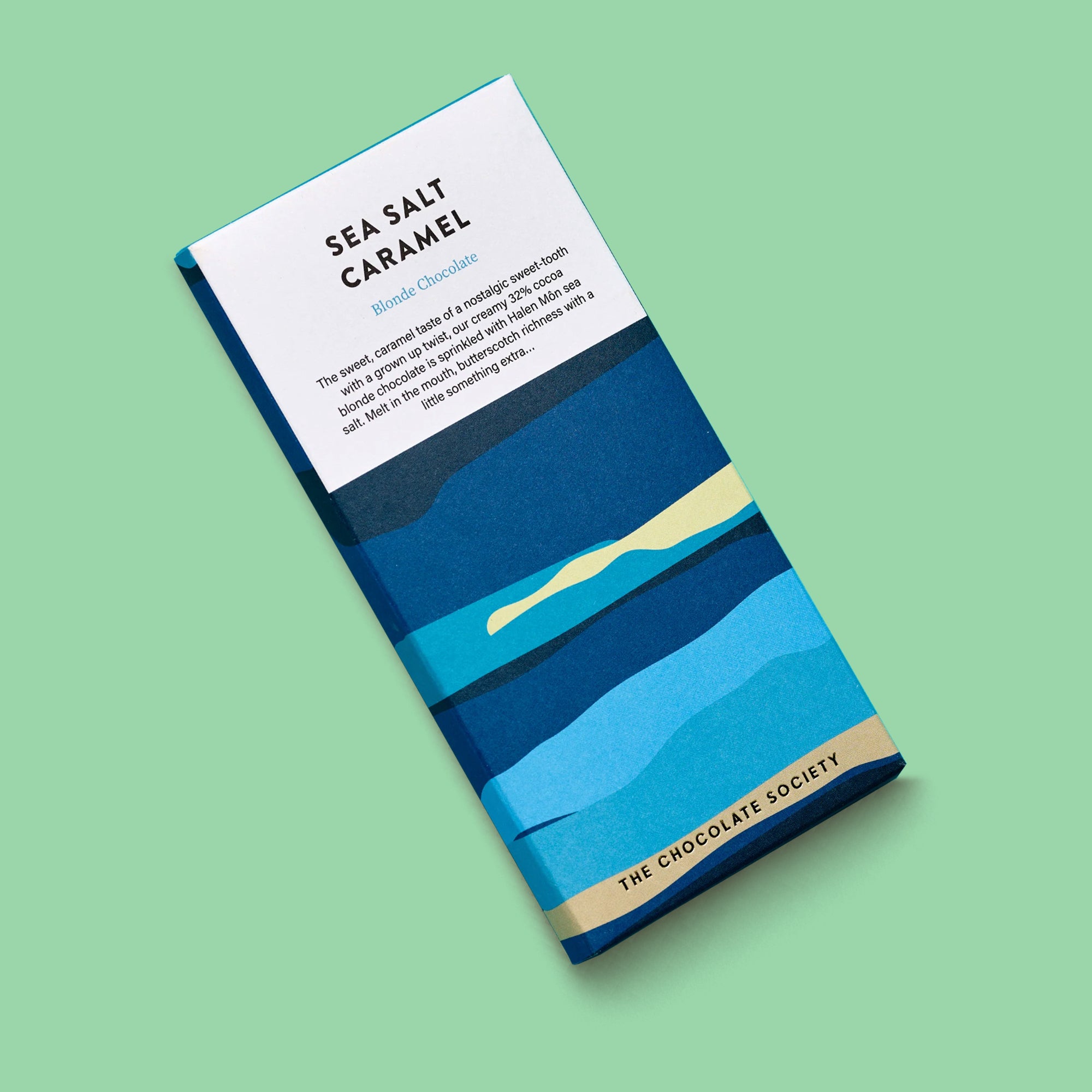 A single chocolate bar package labeled 'SEA SALT CARAMEL' by The Chocolate Society, shown at an angle on a solid mint green background. The wrapper features a stylized blue and navy landscape design with a pale yellow beach and a white upper section containing product text.