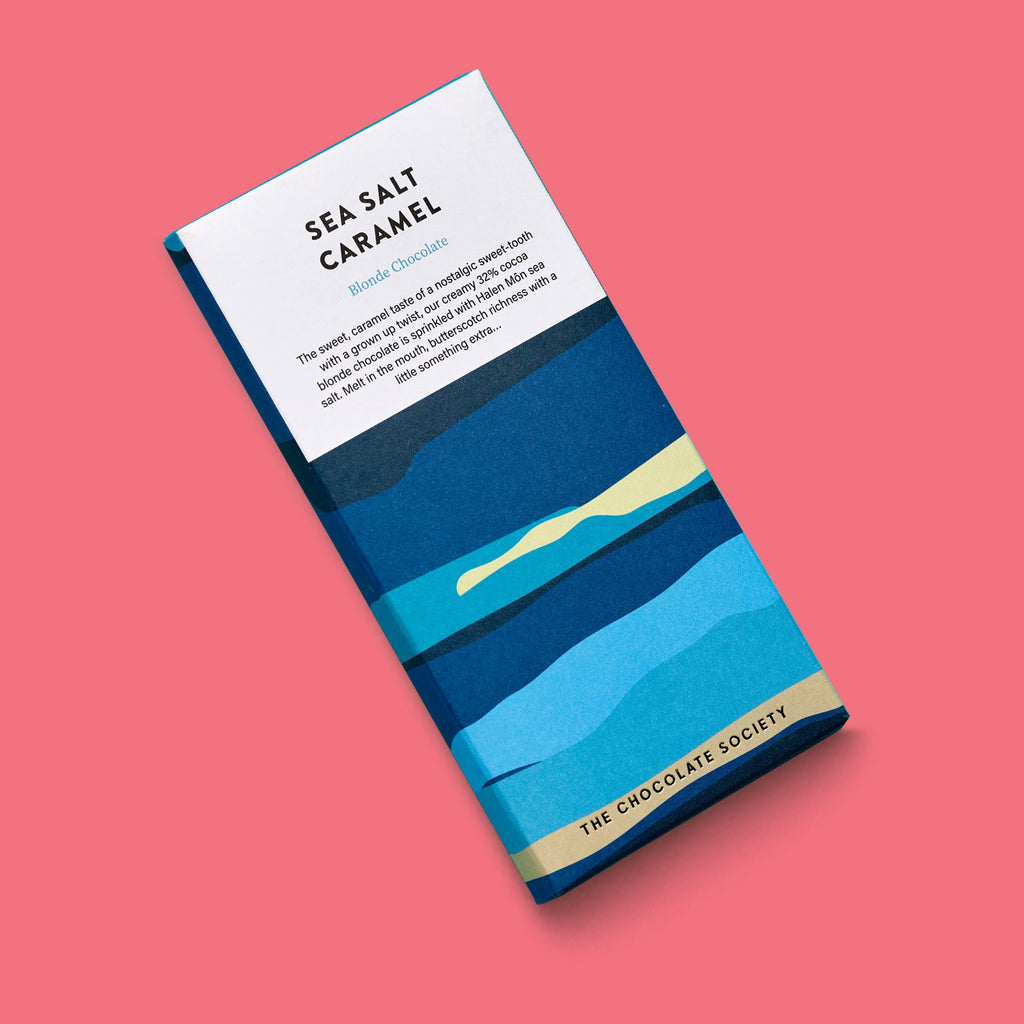 A single chocolate bar package labeled 'SEA SALT CARAMEL' and 'Blonde Chocolate' by The Chocolate Society, shown at an angle on a solid coral pink background. The wrapper features a stylized blue beach landscape with layered sky and sand colors.