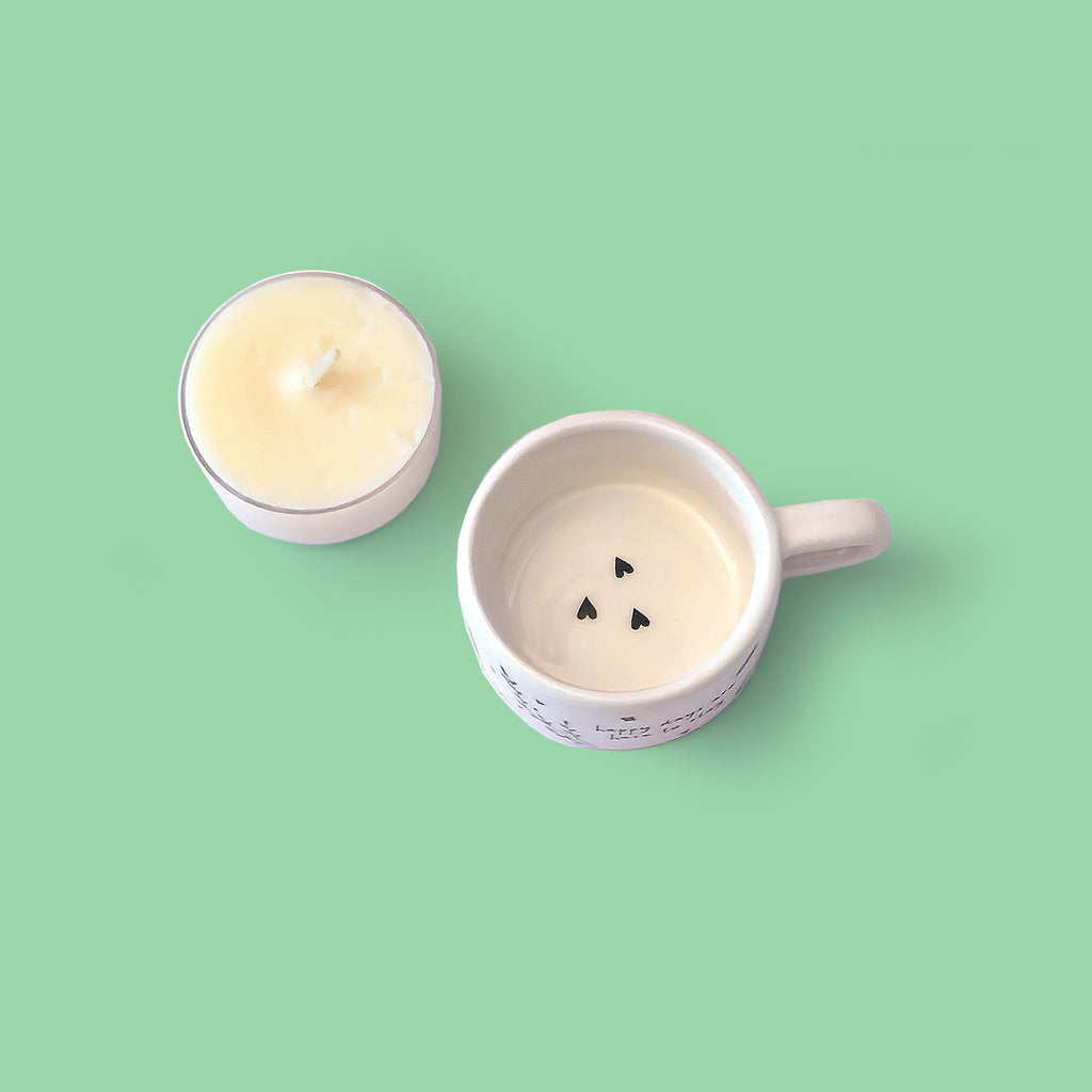 A white ceramic tealight holder and a small tealight candle on a solid mint green background. The tealight holder has three small black love hearts arranged in the bottom.