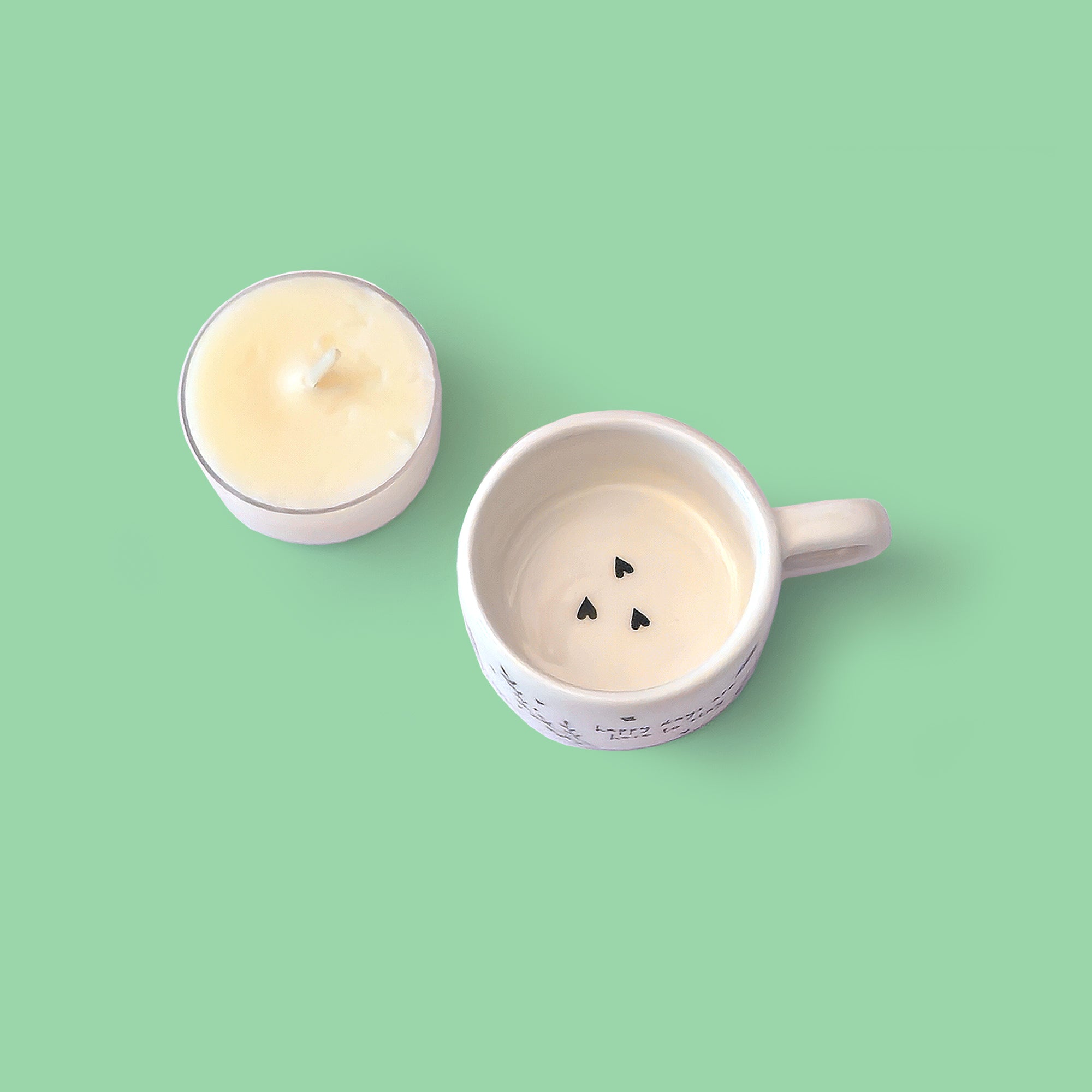 A white ceramic tealight holder and a small tealight candle on a solid mint green background. The tealight holder has three small black love hearts arranged in the bottom.