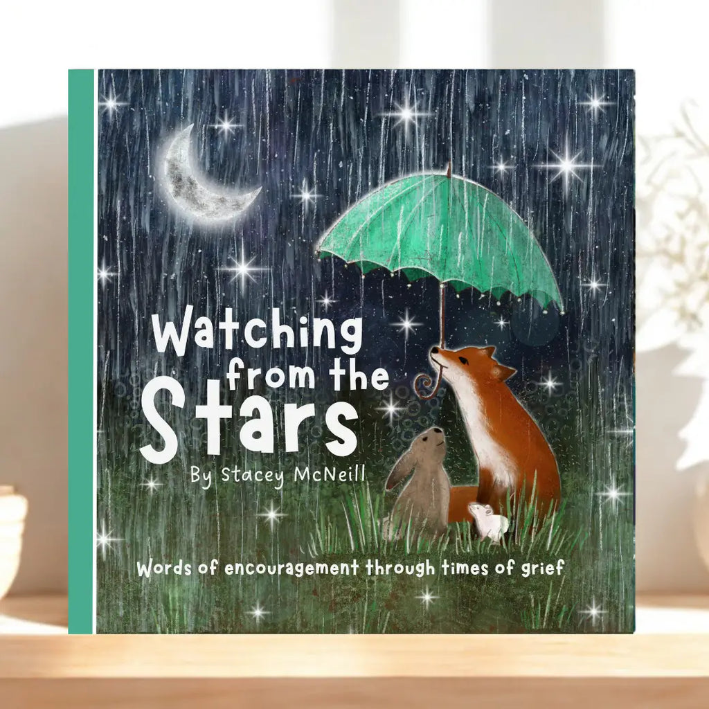 A grief book titled 'Watching from the Stars' by Stacey McNeill, featuring a whimsical illustration of a fox, rabbit, and cat under a green umbrella in the rain at night, displayed on a light wooden surface.