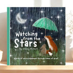 A grief book titled 'Watching from the Stars' by Stacey McNeill, featuring a whimsical illustration of a fox, rabbit, and cat under a green umbrella in the rain at night, displayed on a light wooden surface.