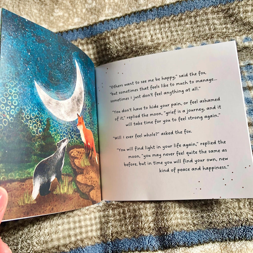 An open grief book lying on a textured blanket, showing a colourful illustration of a fox and moon on one page and several lines of printed text on the other page.