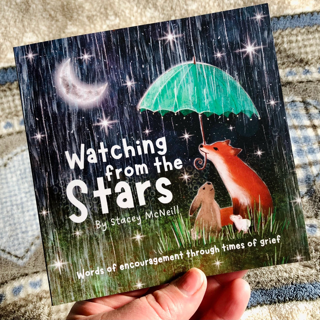 A hand holds a grief book titled 'Watching from the Stars' by Stacey McNeill. The cover features a dark rainy night scene with a crescent moon, stars, a green umbrella, and a fox, rabbit, and puppy under the rain.