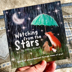 A hand holds a grief book titled 'Watching from the Stars' by Stacey McNeill. The cover features a dark rainy night scene with a crescent moon, stars, a green umbrella, and a fox, rabbit, and puppy under the rain.